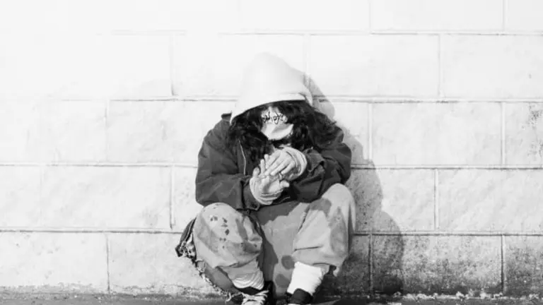 In black and white, they crouch against a wall covering their face
