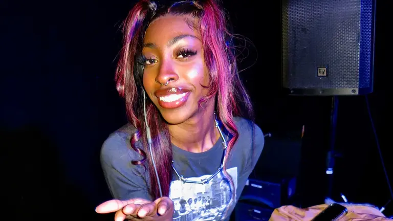 emski wearing a black t-shirt in a dark room, smiling into the camera with the flash on with her hand stretched out to the screen