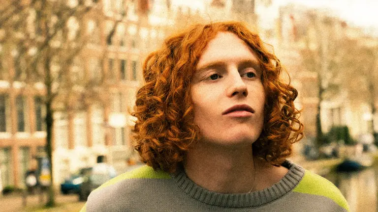 a man with red curly shoulder length hair and a green jumper, outside, looking away from the camera