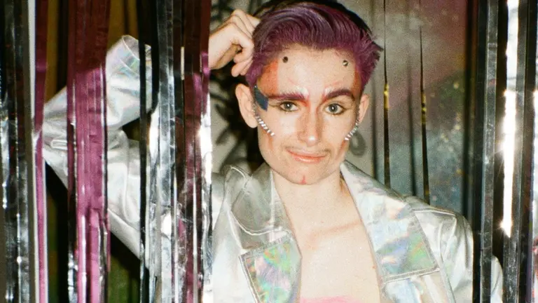 A person leans against a silver wall, with purple hair, an open silver jacket with on t-shirt on
