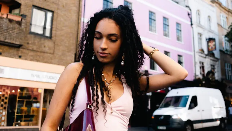 A woman in front of a pink building and white van, with a pink top on, with her hand in her hair and her eyes closed, looking content