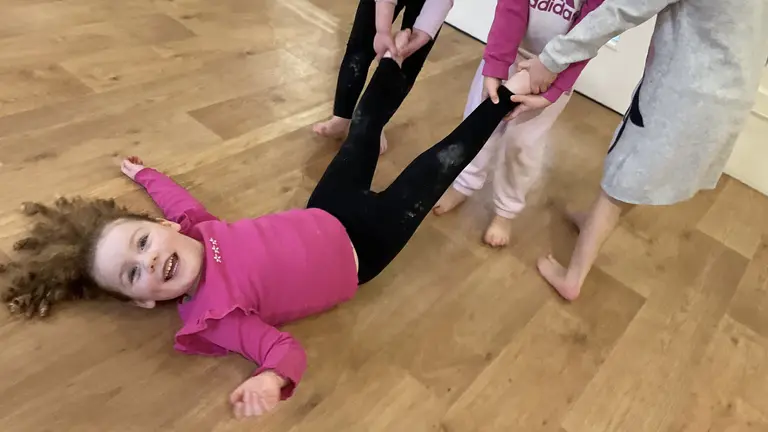 a girl with a pink top and dark leggings being dragged across the floor smiling by three other children, looking at the camera