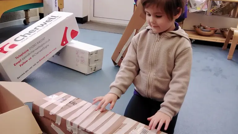 A child stands in front of a cardboard that has been covered in pieces of white masking tape. Their fingers are pressed against the box, almost like they are about to play the piano. 