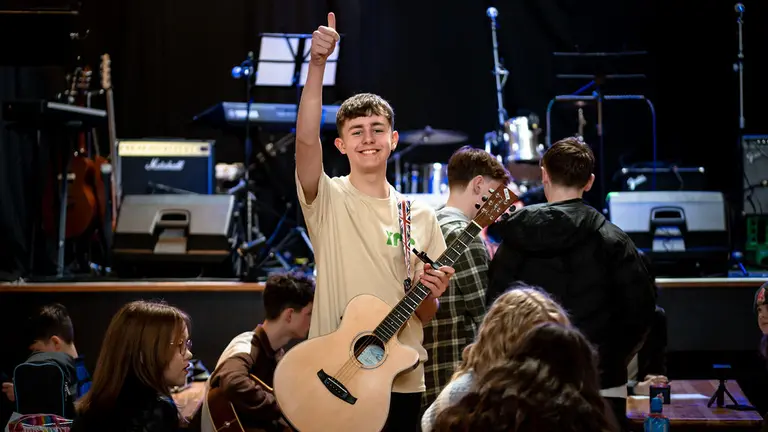 A young boy with an acoustic guitar over his shoulder stands, smiles and gives a big thumbs up to the camera. He is standing in front of a stage with other people around.