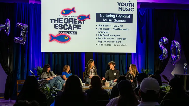 five people sit in chairs on a stage as part of a panel, in front of an audience