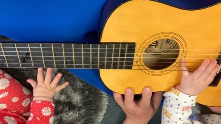 Two toddlers and an adult play with a guitar - we only see the hands. 