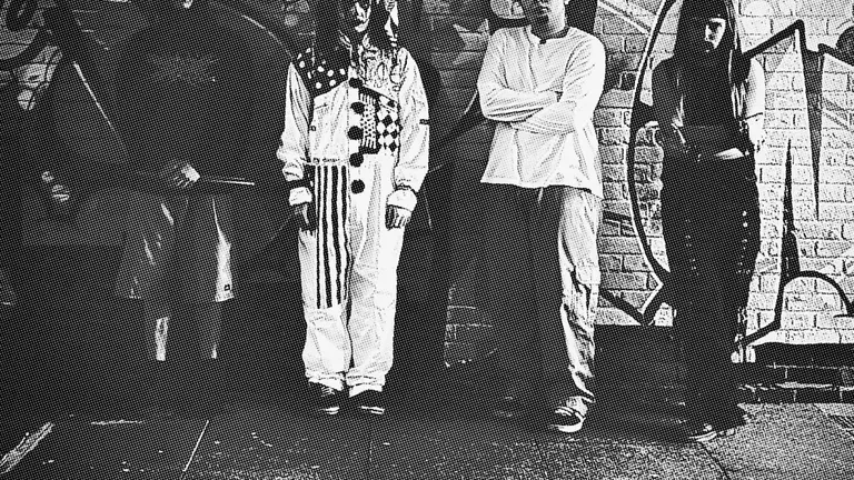 Black and white picture of four people standing against a wall full of graffiti. 