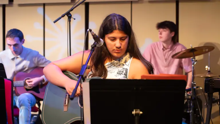 a young person in the foreground playing the guitar, with two other younger people behind, one of drums and one on guitar