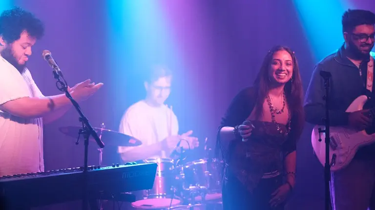 Four people under the lights of a scenario. One is signing, another in the drums, another is playing the guitar and the last one is at the keyboards 