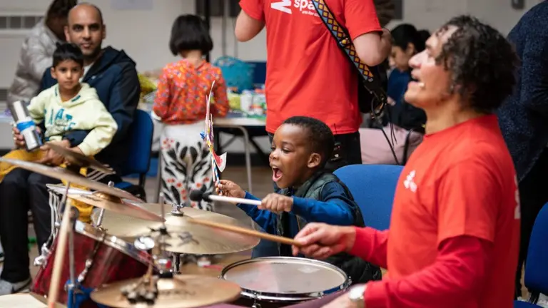 A young child playing drums with an adult. In the background another adult with a child watch the scene. 