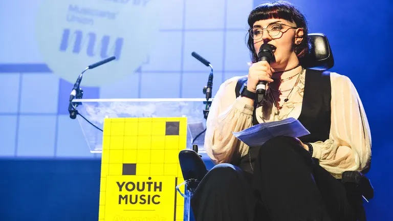 Elizabeth J Birch holding a microphone and giving a speech on stage at the Youth Music Awards