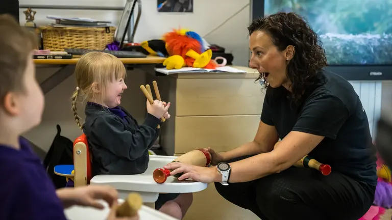 An adult and a child laugh together. The child has pencils in their hands and the adult is kneeing to their level. 