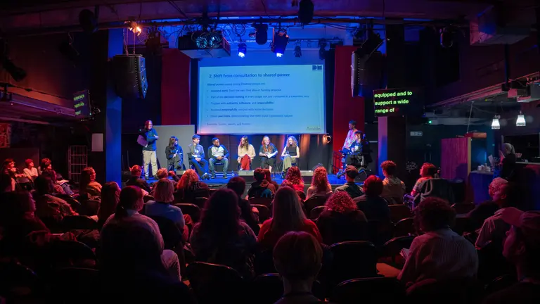Public in front of a scenario. The venue is dark and there's a screen at the front. 