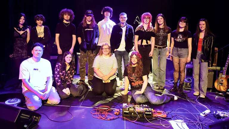14 young people in a scenario. Four are seating and the rest are standing up, all looking at the camera