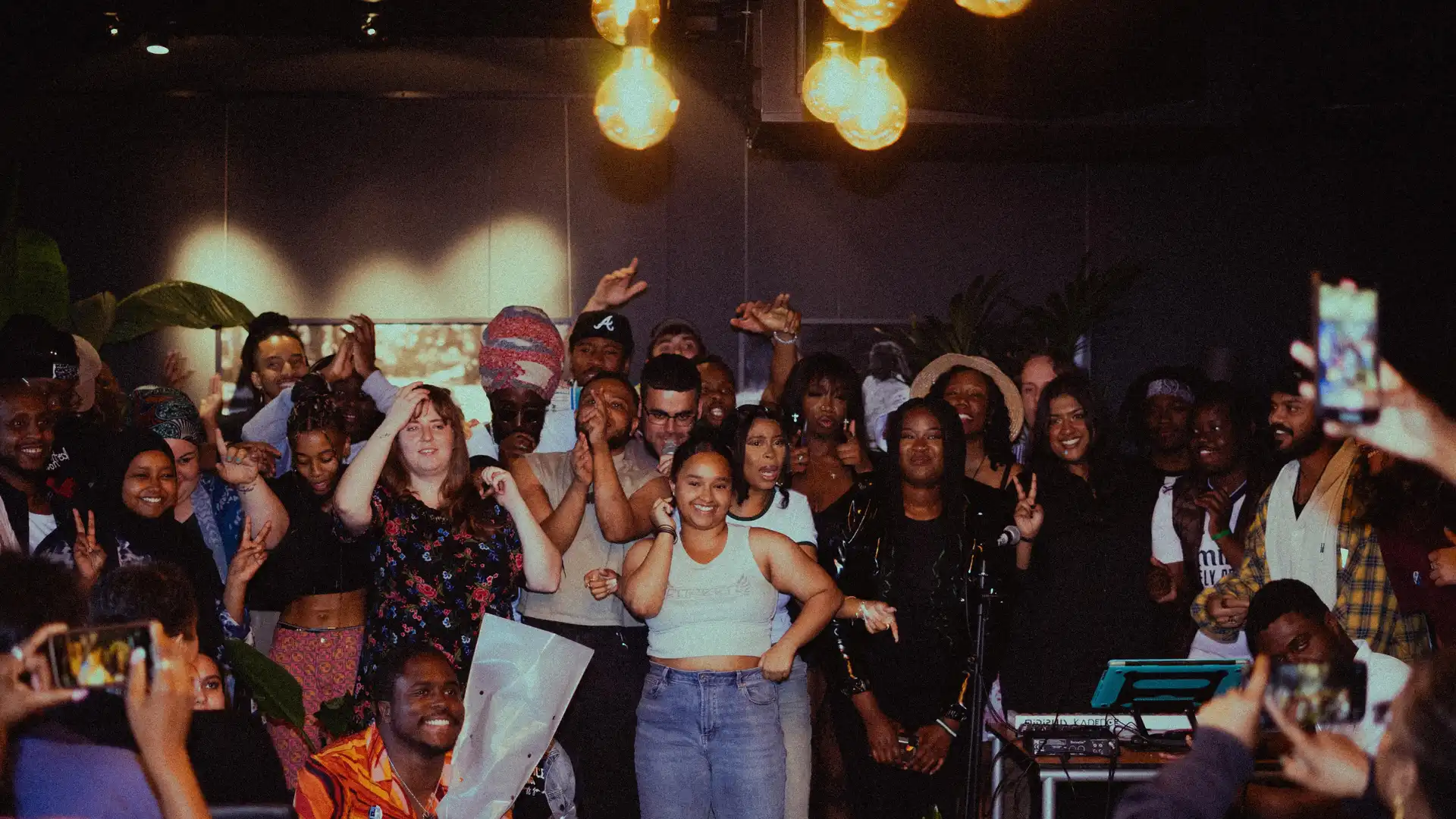 A group of young people at a music project are gathered in a warmly lit room, posing for a group photo. The scene conveys joy and togetherness.