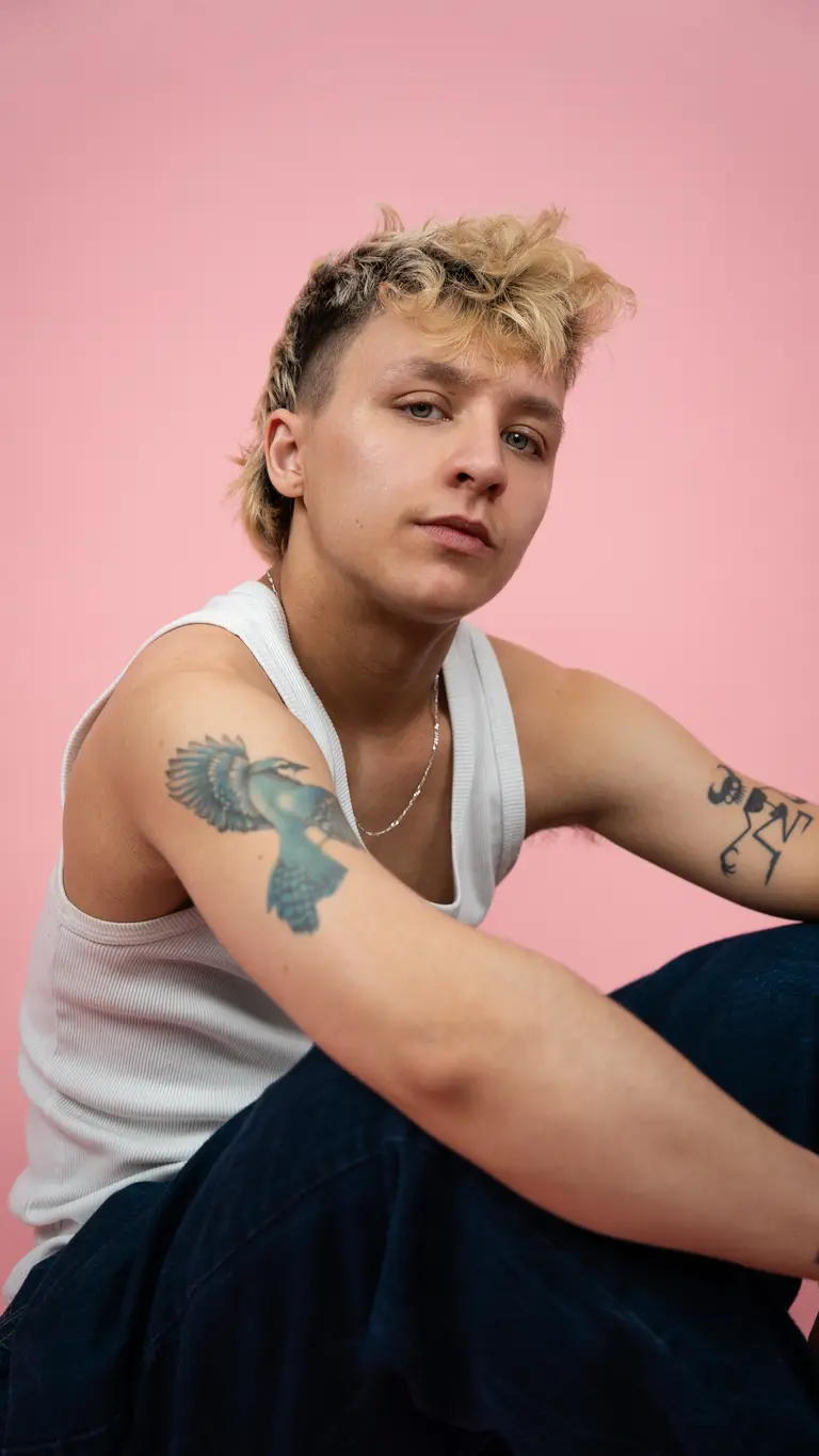 Indy has brown and blonde hair cut into a mullet. he wears a white tank top and shows his arm tattoos