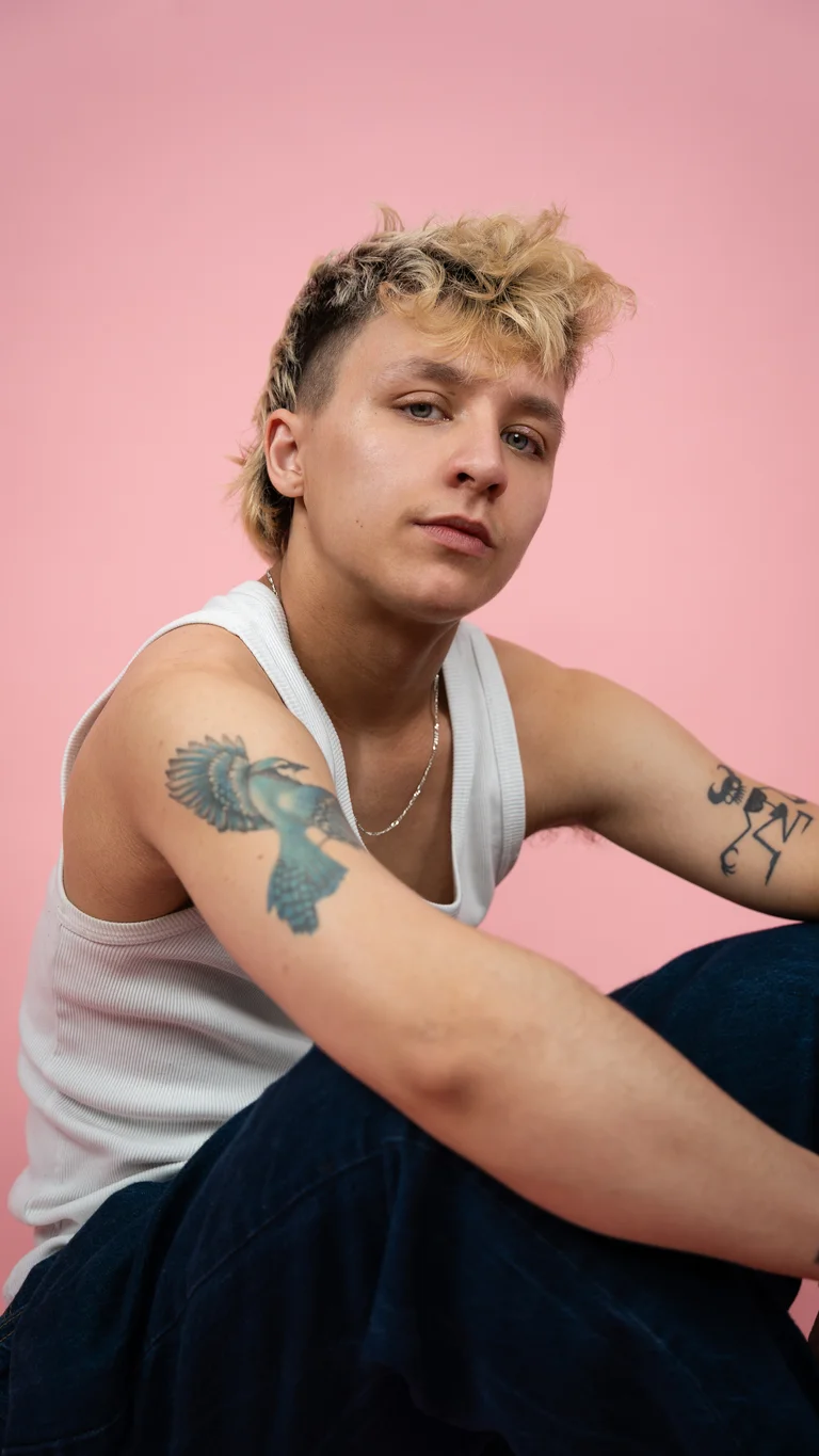 Indy has brown and blonde hair cut into a mullet. he wears a white tank top and shows his arm tattoos