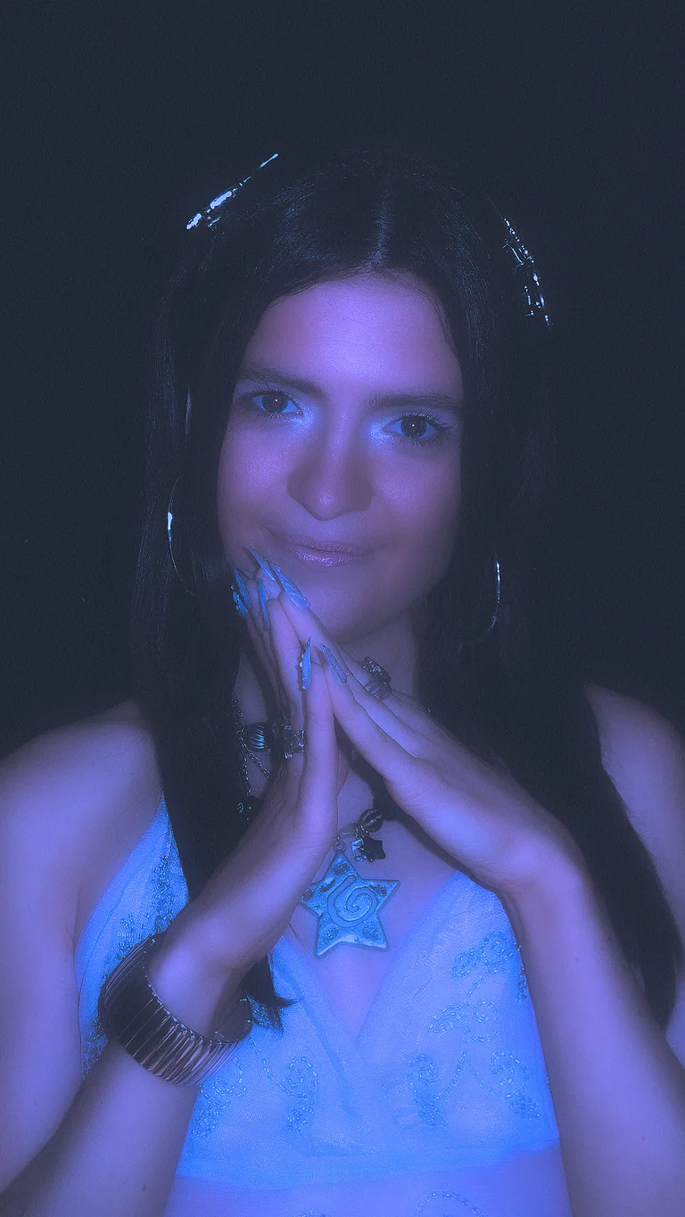 melba holds her hands together by her chin. her eyes are traced in a shimmery blue