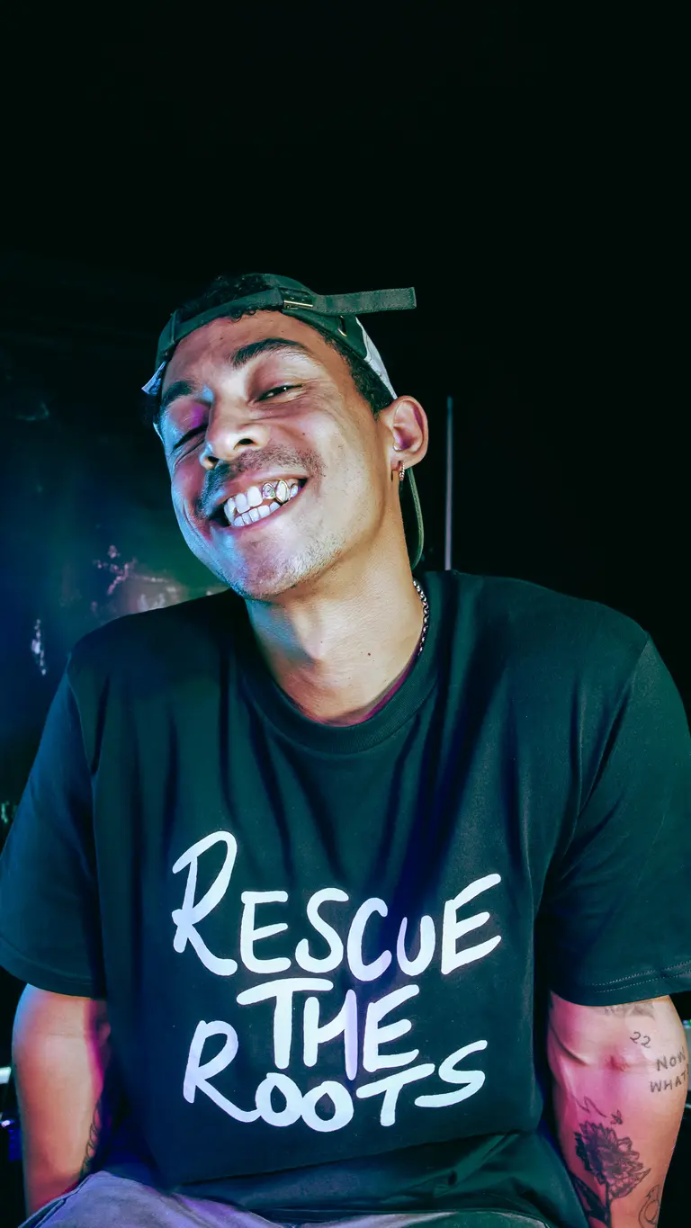 A smiley Jordan Stephens wearing a black t-shirt that says Rescue The Roots. he's wearing a cap. 