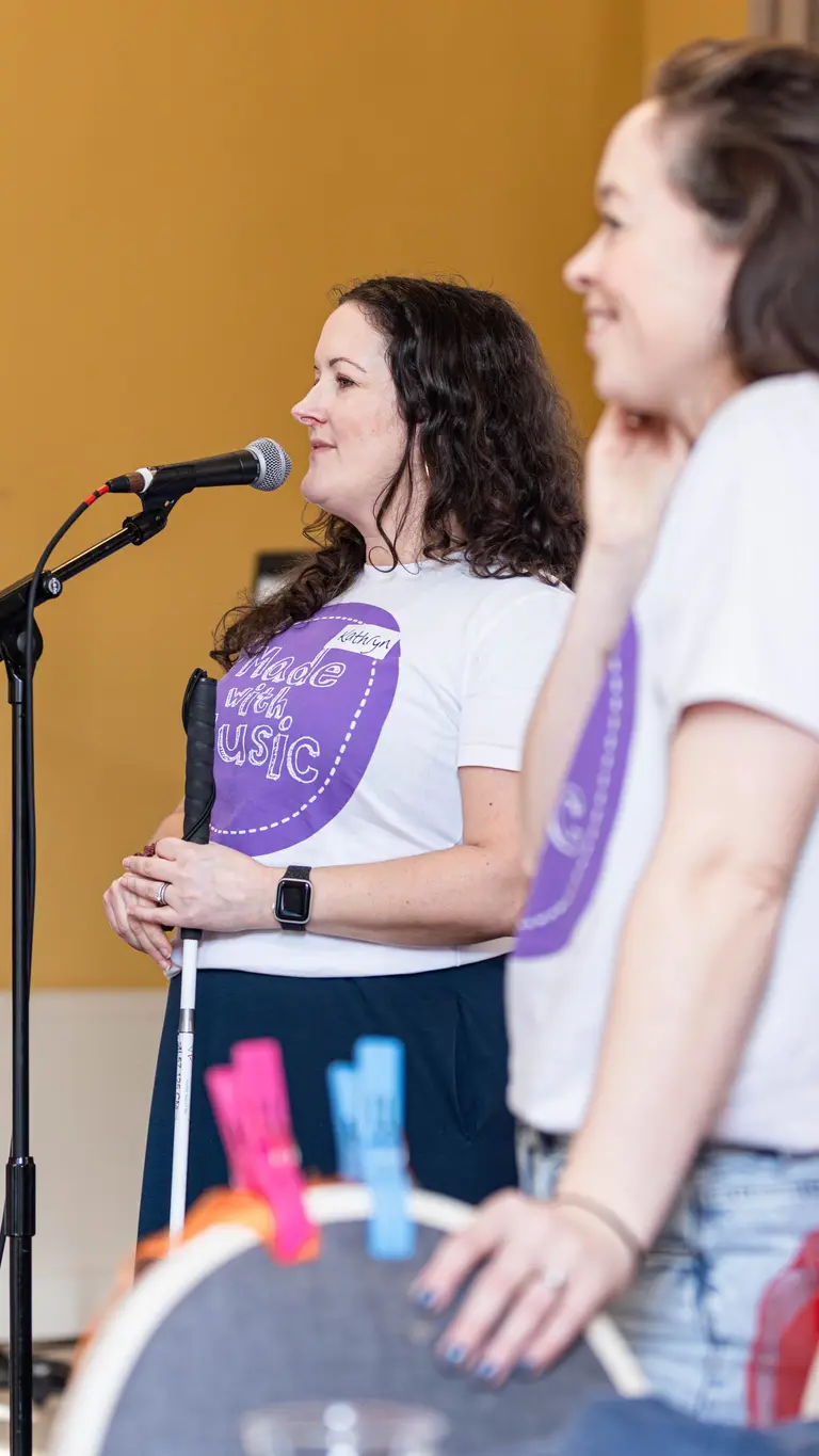 two women with 'made with music' t-shirts. One is stood in front of a microphone talking, the other, in the foreground, is leaning on a chair and smiling