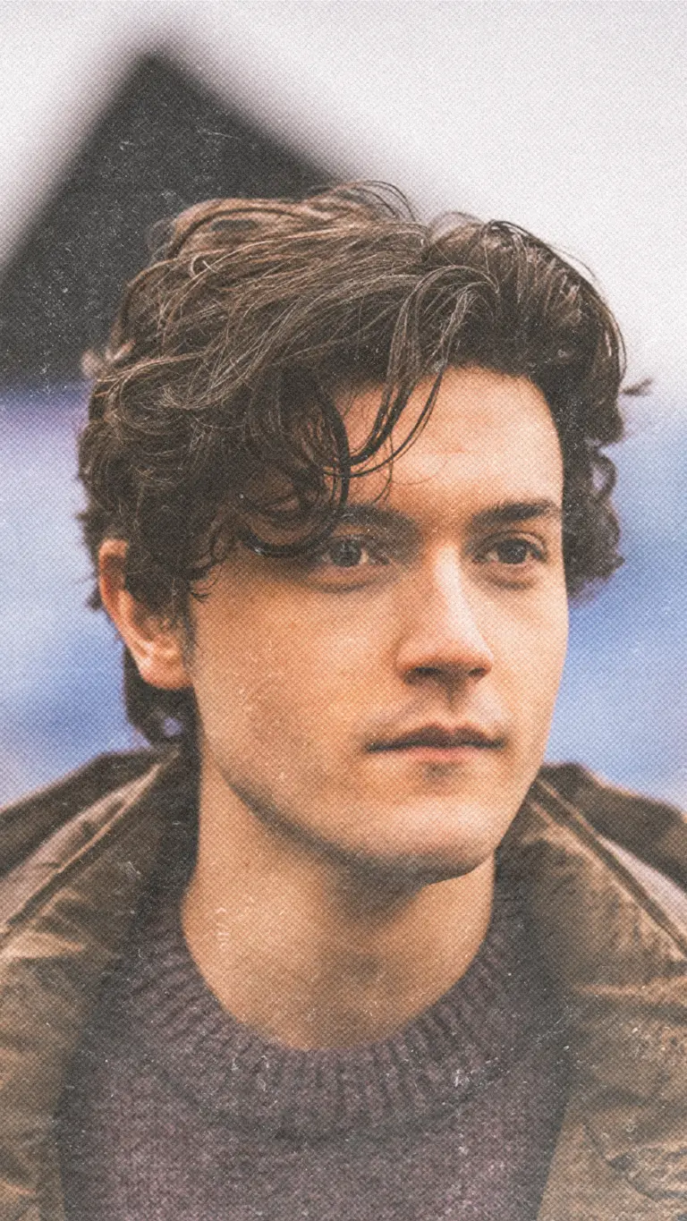 a young man with dark curly cropped hair wears a brown jacket and purple knitted jumper