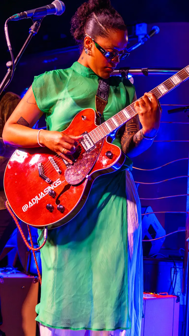 adjua wears a green dress, sunglasses, and holds a red electric guitar