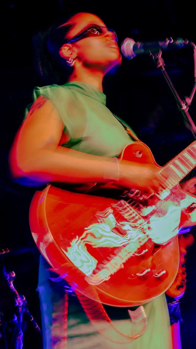 adjua wears a green dress, sunglasses, and holds a red electric guitar