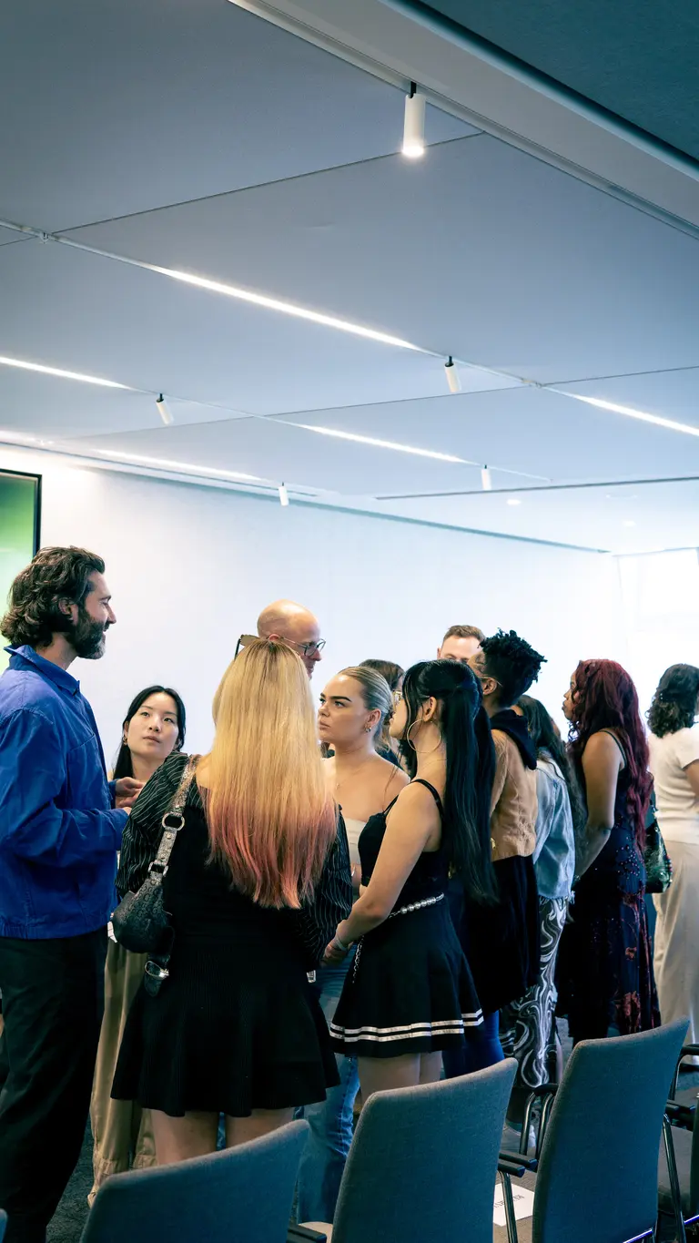 a group of young people listen to a man talking. they are standing up in a room with chairs behind