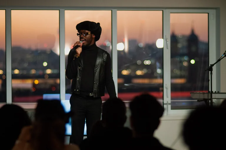 a person wearing glasses and an all-black outfit stands on stage, speaking into a microphone, with a sunset cityscape behind them
