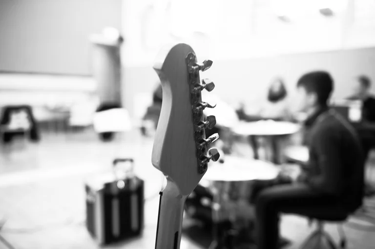 a guitar head is in focus, a music room with people in the background