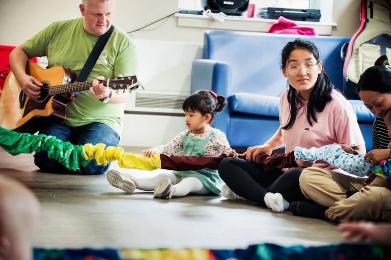 Image of a young child sat on the floor, with adults, singing and with a coloured rope on their lap