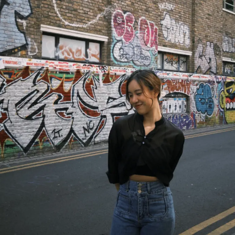 TIffy standing with her arms behind her back and smiling. She is wearing a black button shirt and blue jeans, on a street with a lot of graffiti on the walls