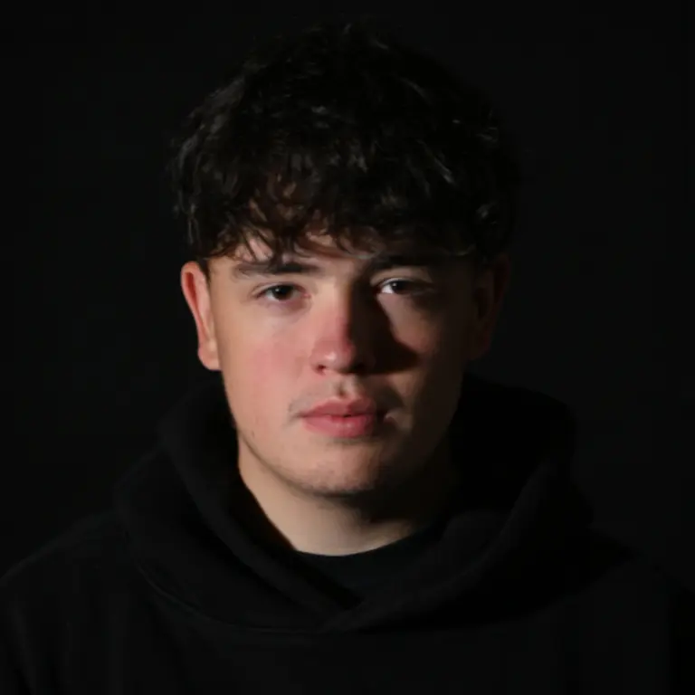 Jack, in a dark image where only his face is lit, is looking straight at the camera with a neutral expression. His shirt, hair and background is all dark. 