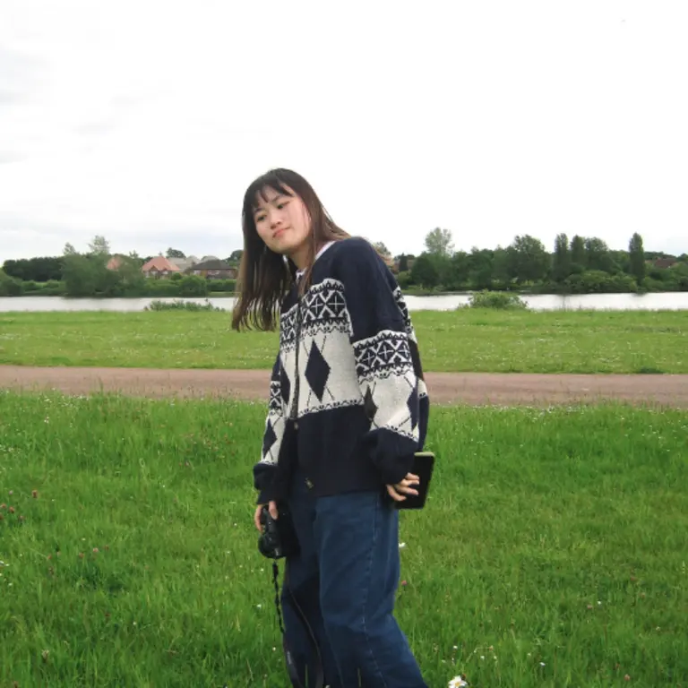 Katie wearing blue jeans and a patterned fleece, leaning away from the camera with her eyes closed. She is in a field with a track and river behind her, it is summer and green
