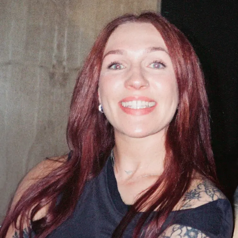 Kate smiling in front of a flash camera, wearing a black top with straight auburn hair and a small tattoo on her left shoulder. 