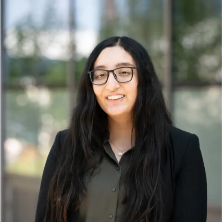 Isha smiling outside of a glass building, wearing glasses. She is wearing formal work clothes, with a black jacket and smiling