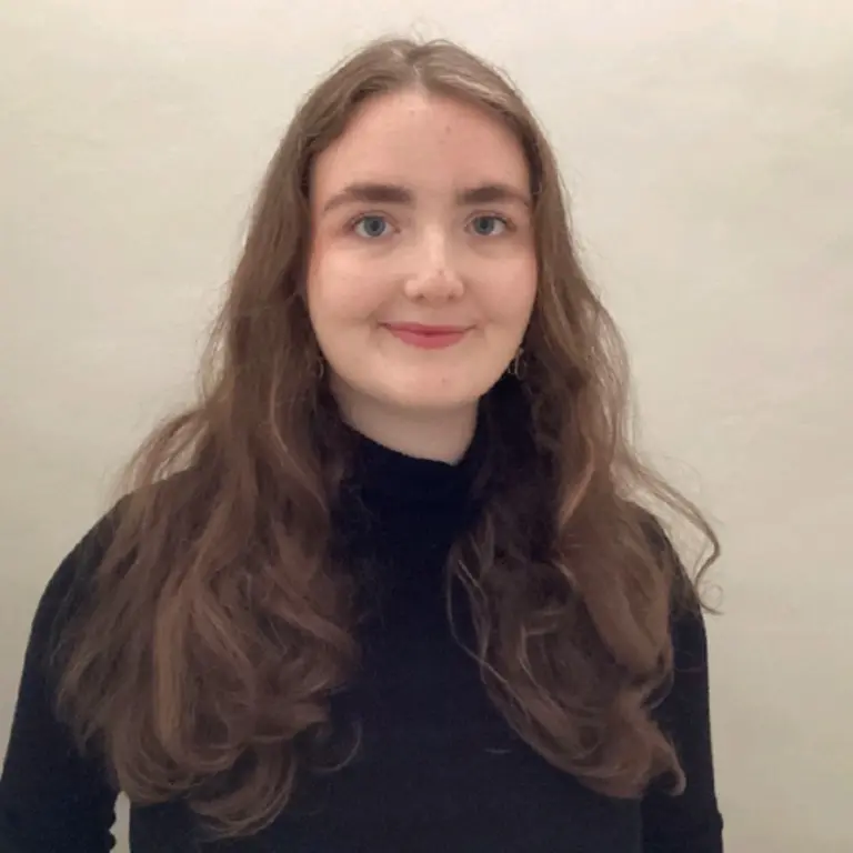Isabel smiling in front of a beige/cream wall. She is wearing a high necked black shirt and smiling, with hair falling over both of her shoulders