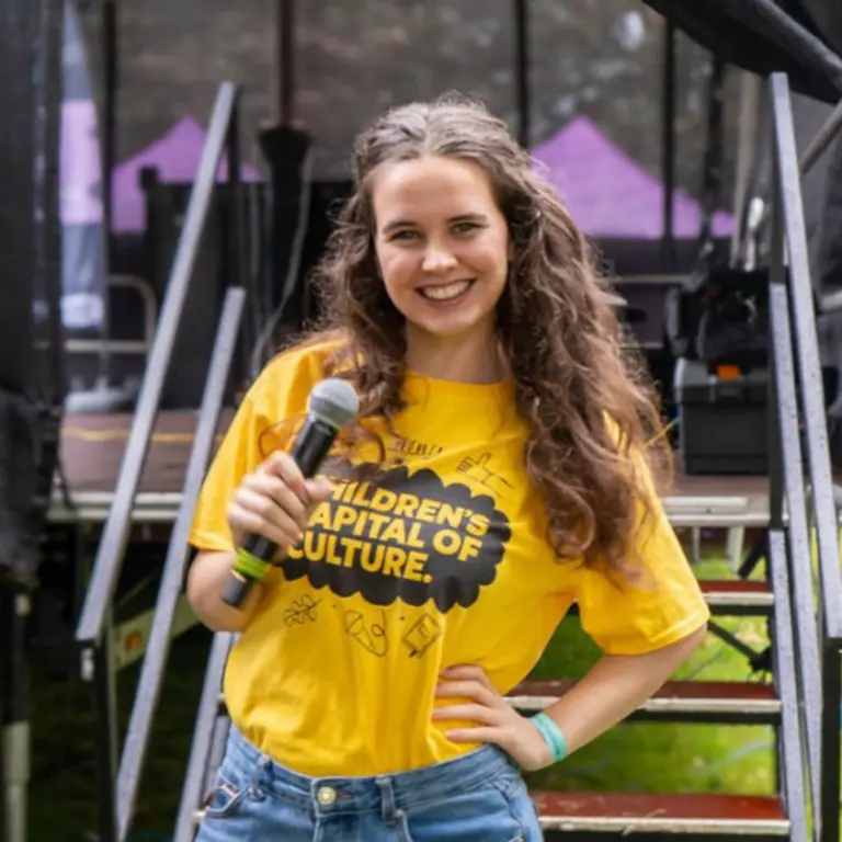 Grace smiling and posing, with her right hand holding a mic, her left hand on her hip. She is smiling with long curly hair, wearing a yellow T-Shirt and blue jeans, behind a stage