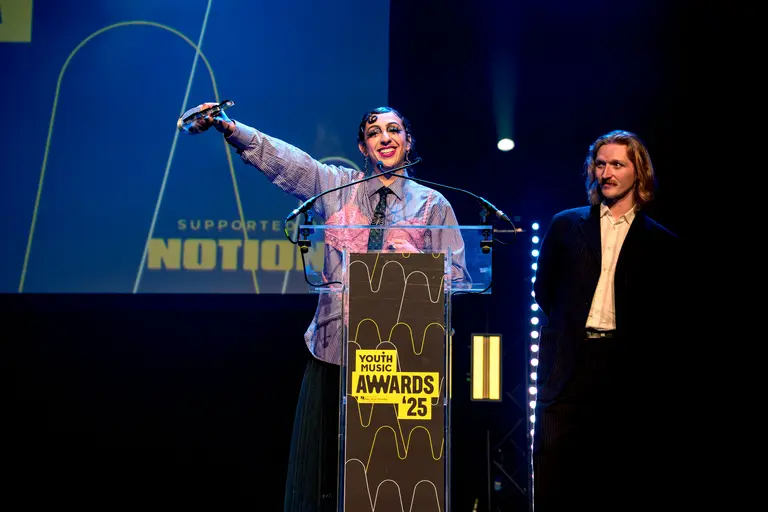 Rivkala on stage at the youth music awards 2025 accepting her award and lifting it in the air to the crowd. Her bandmate is behind her smiling