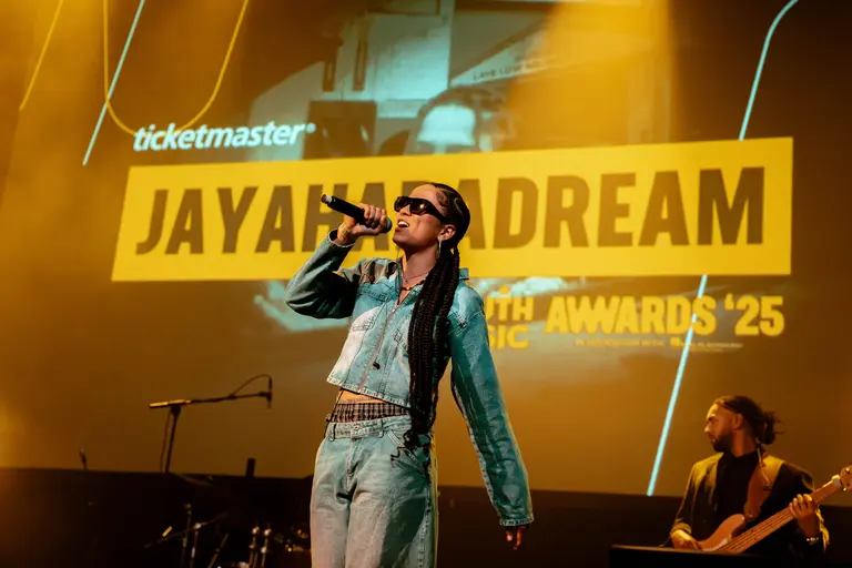 jayahadadream performing in a denim top and jeans. She's on stage at the YMAs 2025, performing with sunglasses on in front of a large screen with her name on it, singing into the mic