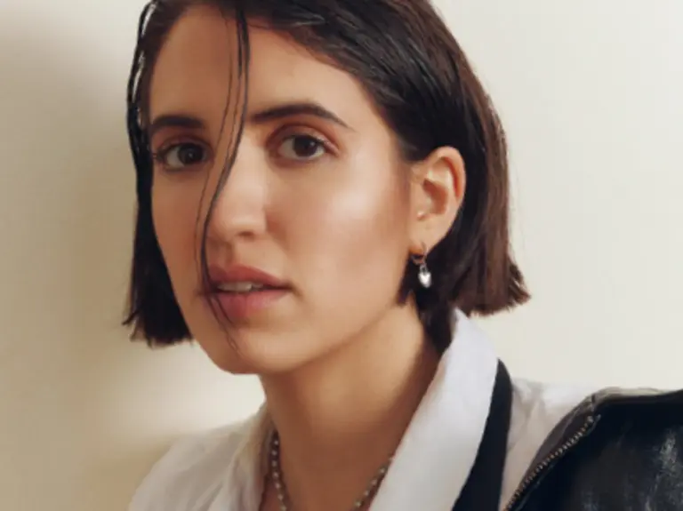 Face of a person looking at a camera, with short hair, a string on hair over their face and a white shirt. 