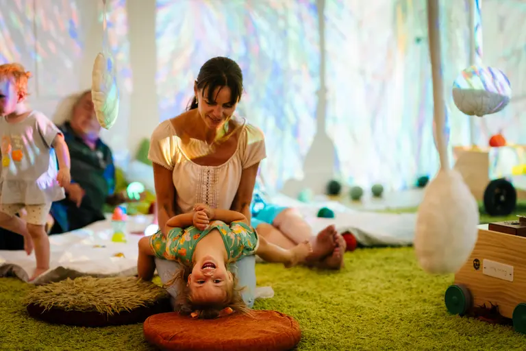 An adult and a young child play together during an early years creative session at Magic Acorns, surrounded by soft cushions, hanging sensory objects and colourful projected light patterns.