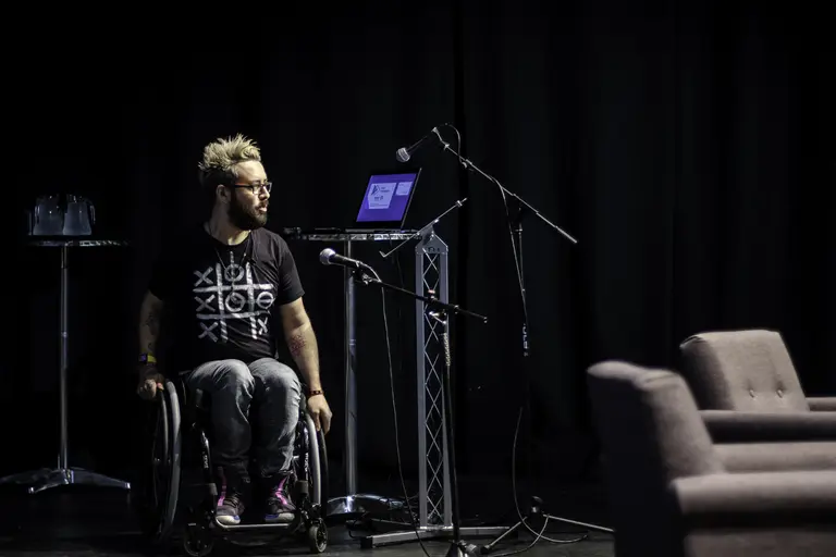 A person in a wheelchair sits on a stage near a podium with a laptop and several microphones. The background is dark, and there are chairs and a small table with water glasses arranged around the stage.
