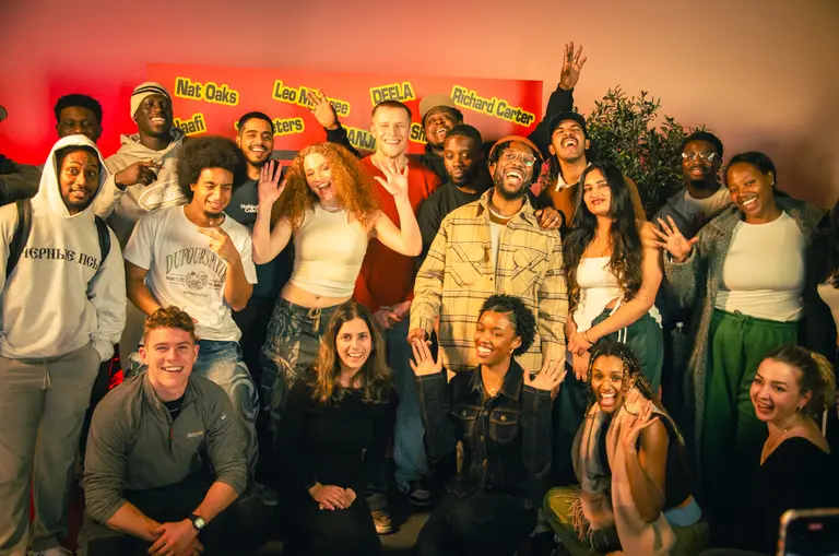Group of young creatives gathered at the Ones To Watch Writing Camp 2025, standing and sitting together in a warmly lit room. Behind them is a bright red backdrop with green and yellow name cards displaying artist names such as Nat Oaks, Leo, DEELA, and Richard Carter. Some participants are raising their hands, and the atmosphere appears lively and collaborative.