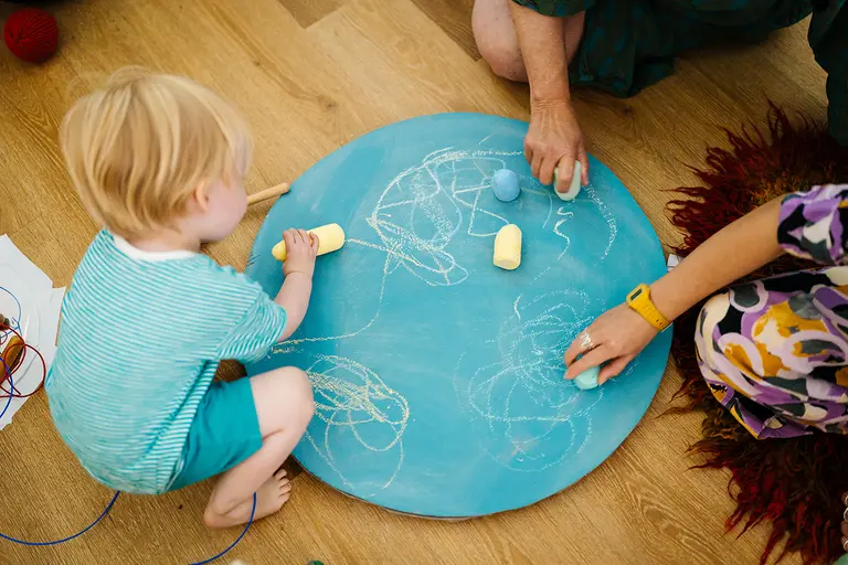 child drawing with chalk on a turqoise board on the floor, with two adult hands also drawing