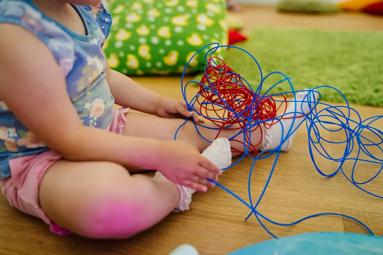 A young child sits on the floor playing with tangled blue and red sensory cords.