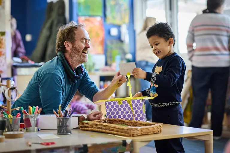 A child explores a ribbon inside a patterned box as an adult supports the activity. Art supplies and colourful classroom displays surround them, creating a playful early‑years learning environment.