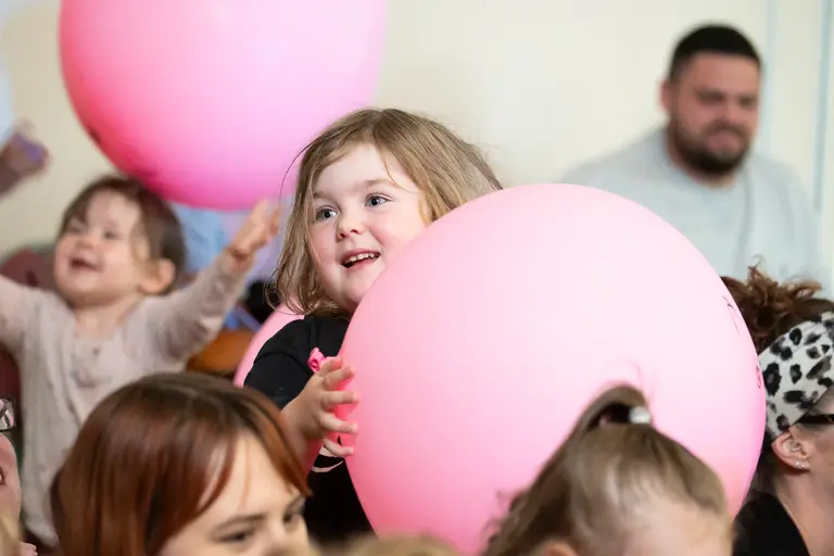 An early years child holding a large pink balloon and smiling among other children holding balloons