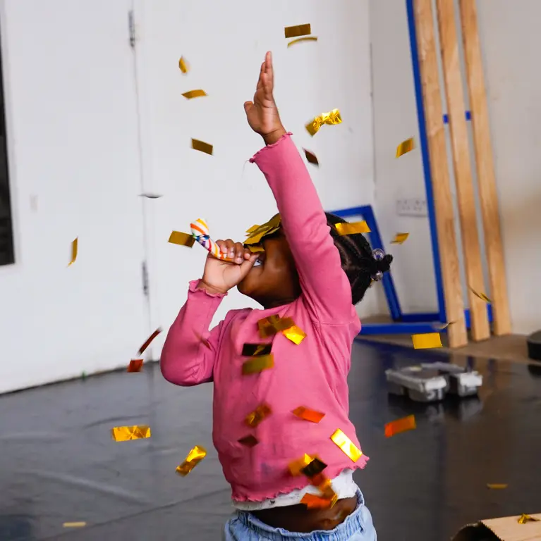 an early years girl with an arm in the air and the other to her mouth while confetti falls on them 