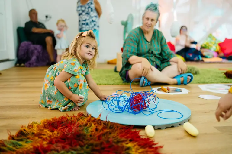 An early years session, with a child on their knees looking and smiling off screen playing with different coloured string. In the background are other children and adults playing